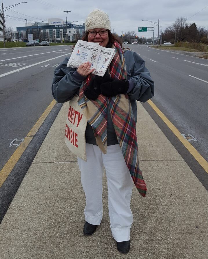 Charity newsie on Drive Day standing on street holding up newspaper