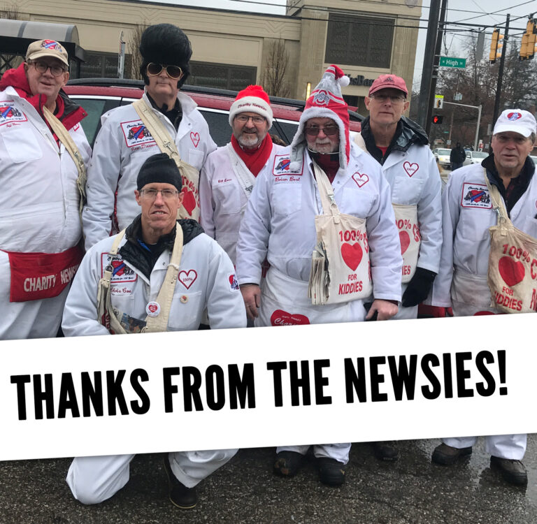 Group of Charity Newsies in white jumpsuits on Drive Day with newspapers with sign saying "Thanks from the Newsies!"